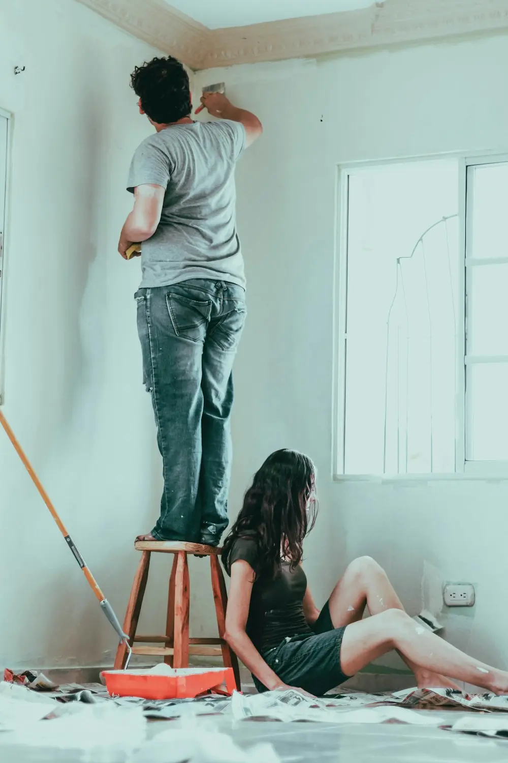 Férfi sámlin állva festi a falat, mellette egy lány ül